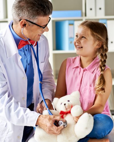 Joyful girl looking at her doctor examining teddy bear in hospital